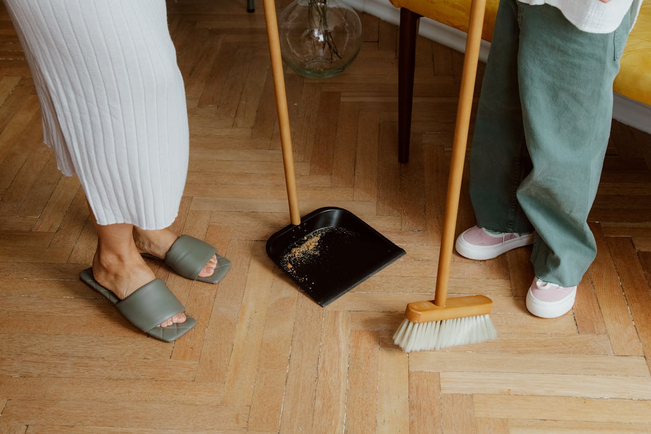 A mother and child cleaning the floor with a broom and dustpan indoors.